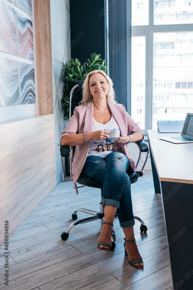 Obraz premium An adult business woman is sitting in an armchair in her office with a cup of coffee in her hands and looking at camera