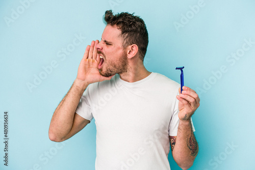 Young caucasian man shaving his beard isolated on blue background shouting and holding palm near opened mouth.