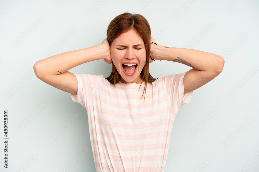 Young English woman isolated on blue background covering ears with hands trying not to hear too loud sound. Young English woman isolated on blue background covering ears with hands trying not to hear too loud sound.
