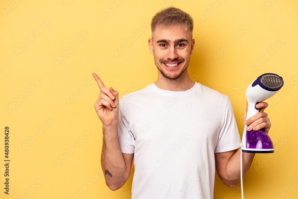 Young caucasian man holding an iron isolated on yellow background smiling and pointing aside, showing something at blank space. Young caucasian man holding an iron isolated on yellow background smiling and pointing aside, showing something at blank space.