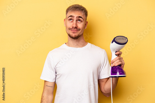 Young caucasian man holding an iron isolated on yellow background dreaming of achieving goals and purposes