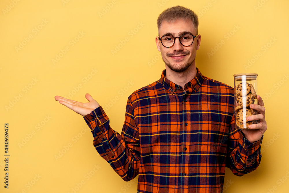 Young caucasian man holding cookies jar isolated on yellow background showing a copy space on a palm and holding another hand on waist. Young caucasian man holding cookies jar isolated on yellow background showing a copy space on a palm and holding another hand on waist.