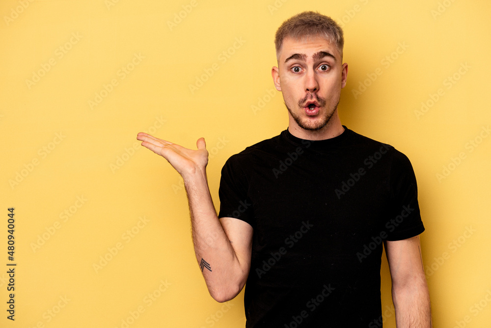 Young caucasian man isolated on yellow background impressed holding copy space on palm. Young caucasian man isolated on yellow background impressed holding copy space on palm.
