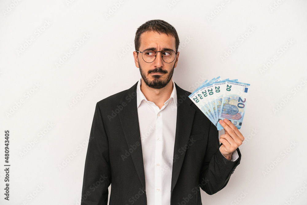 Young caucasian business man holding banknotes isolated on white background confused, feels doubtful and unsure. Young caucasian business man holding banknotes isolated on white background confused, feels doubtful and unsure.