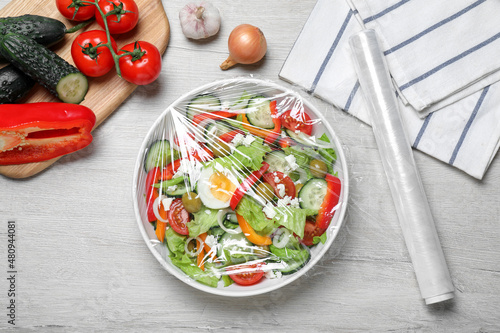 Obraz na plátne Bowl of fresh salad with plastic food wrap on white wooden table, flat lay
