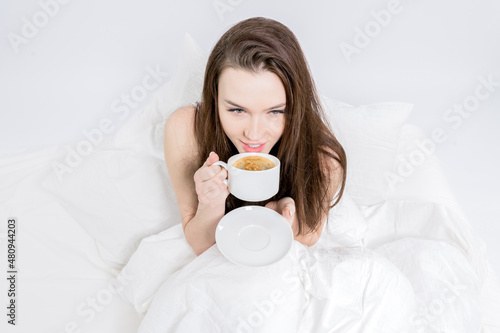 A woman sits in bed and takes a sip of aromatic coffee. Tasty breakfast. The brunette is holding a cup with a hot drink. White bedroom. Good morning