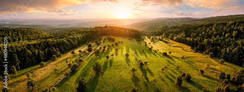 Aerial landscape panorama after sunrise: gorgeous scenery with the sun, trees on meadows in warm light casting long shadows, surrounded by forests