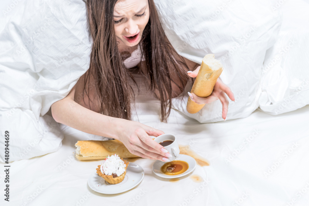 The girl is indignant, spilled coffee on the bed. Breakfast. The brunette lies in bed, next to her is a cupcake, a baguette in her hand and a cup The girl is indignant, spilled coffee on the bed. Breakfast. The brunette lies in bed, next to her is a cupcake, a baguette in her hand and a cup