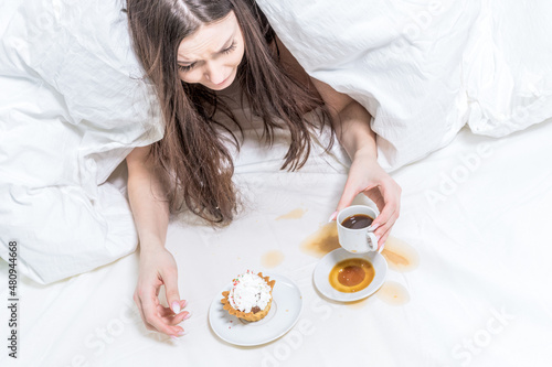 The girl is indignant, spilled coffee on the bed. The brunette lies in bed, next to her is a cupcake, a baguette in her hand and a cup. Breakfast