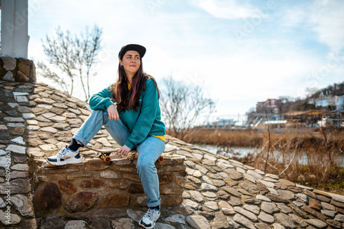 A young smiling Caucasian woman sits on a skateboard and looks away. In the background, the blue sky with clouds and stone paving stones. Copy space. Concept of sports lifestyle and street culture