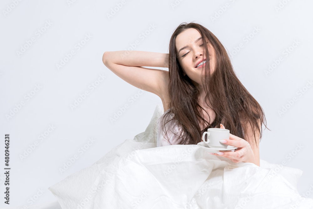 The brunette is holding a cup with a hot drink. Good morning. A woman is sitting in bed pulling herself up with a cup of coffee in her hands. Tasty breakfast. White bedroom The brunette is holding a cup with a hot drink. Good morning. A woman is sitting in bed pulling herself up with a cup of coffee in her hands. Tasty breakfast. White bedroom