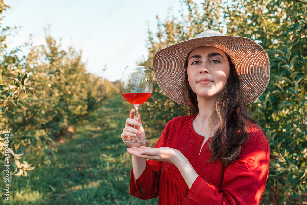 Wine tasting and relaxing in the garden. Young beautiful brunette full face in a red dress and hat with a glass of wine. There are fruit trees in the background.