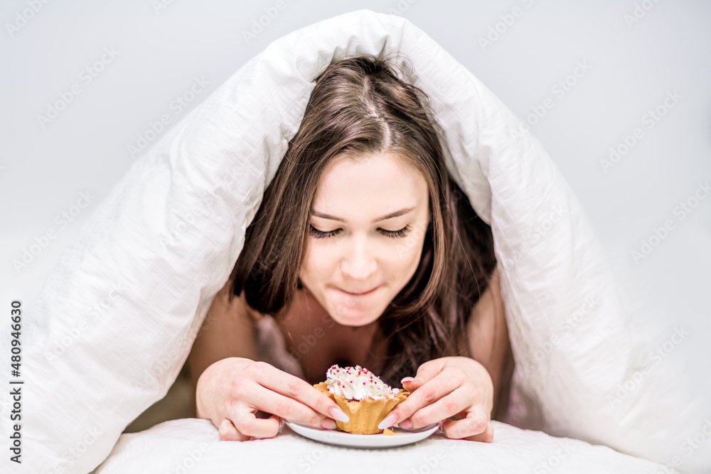A cute woman looks with appetite at a cupcake lying on the bed under the covers. Breakfast in bed. Dessert. Calories A cute woman looks with appetite at a cupcake lying on the bed under the covers. Breakfast in bed. Dessert. Calories