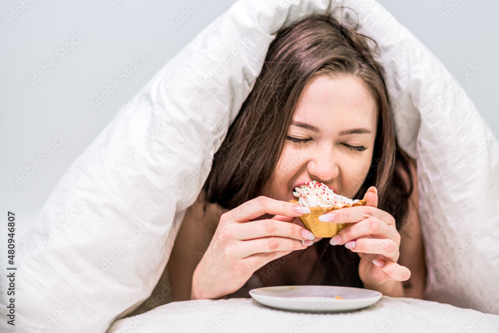 Breakfast in bed. Calories. Dessert. A cute woman with appetite eats a cupcake lying on the bed under the covers Breakfast in bed. Calories. Dessert. A cute woman with appetite eats a cupcake lying on the bed under the covers