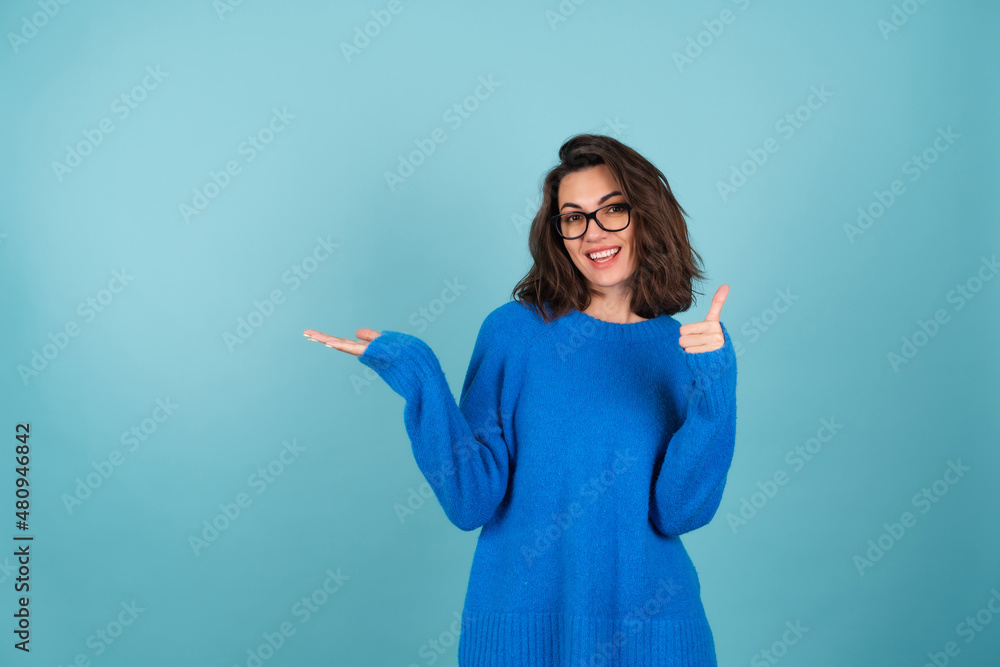 A woman in a blue knitted sweater, curly short hair, glasses on her eyes, smiles cheerfully, points to an empty space, holds in her palm A woman in a blue knitted sweater, curly short hair, glasses on her eyes, smiles cheerfully, points to an empty space, holds in her palm