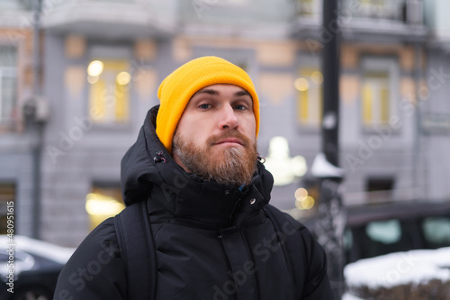 A young man with a red beard in an orange hat in the winter on a proud street in the evening looks at the camera