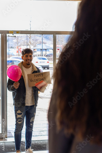 vertical portrait of young latin man smiling giving a surprise with a happy valentine sign, a balloon and a gift to his partner at the door of his front door.