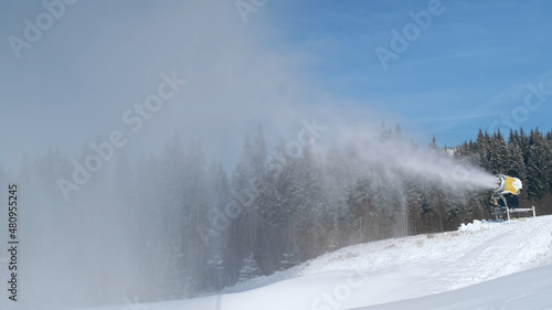 Snow cannon blows artificial snow on mountain slope, background of forest, blue sky in ski resort on sunny day. Snowgun makes snow of water in cold winter. Snowmaker sprays water on mountainside