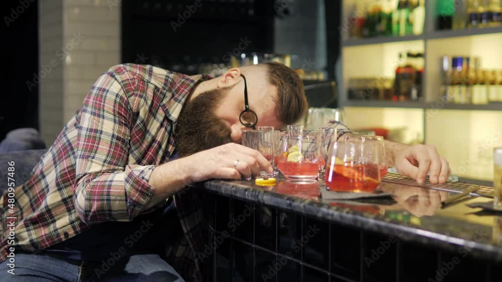 A very drunk young man is lying with his head on a table in a bar. A drunk man in depression sleeps, wakes up and drinks a glass. There are a lot of empty glasses on the table next to the client. A very drunk young man is lying with his head on a table in a bar. A drunk man in depression sleeps, wakes up and drinks a glass. There are a lot of empty glasses on the table next to the client.