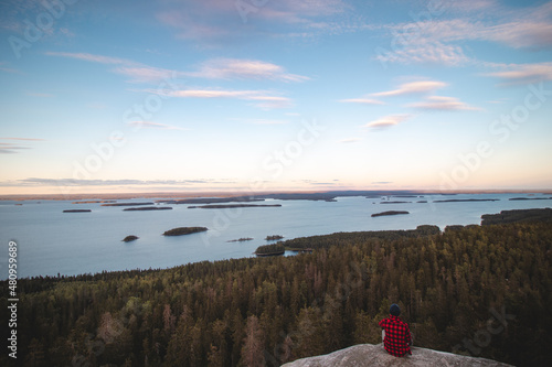 View of an adventurer in red and black shirt sitting on the edge of a cliff watching Lake Pielinen in Koli National Park in eastern Finland. Sunset over Lake Suomi. Magical view of green biodiversity photo