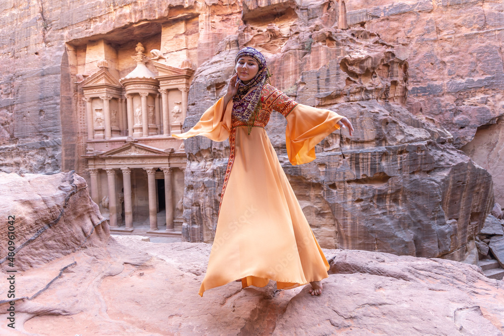 woman dressed in traditional clothes and headscarf staying at the top of rock on the background is Petra palace Jordan