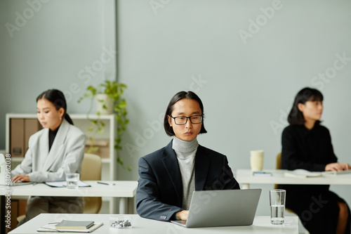 Minimal portrait of Asian businessman using laptop with female colleagues in background, copy space