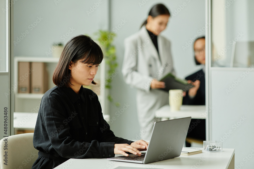 Side view portrait of young Asian woman using laptop at desk in office cubicle, copy space Side view portrait of young Asian woman using laptop at desk in office cubicle, copy space