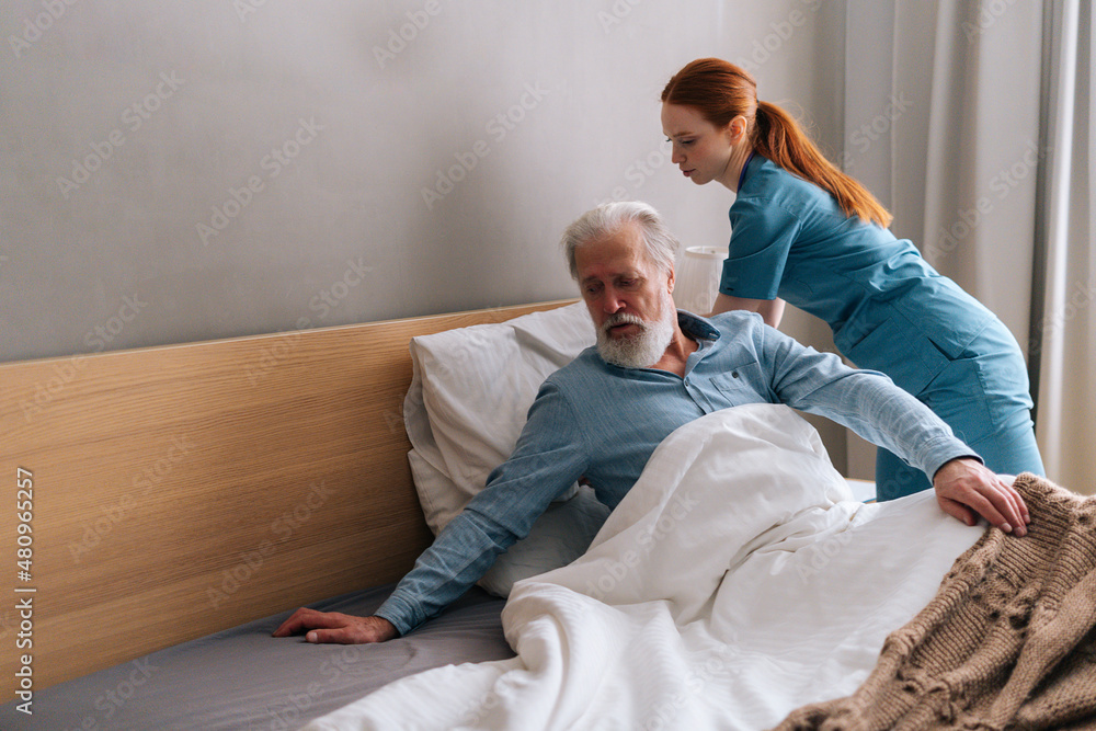Positive young female home caregiver helping senior adult male patient to lying on bed in bedroom at nursing home. Professional nurse helping old man to lying on bed in luxury hospital room. Positive young female home caregiver helping senior adult male patient to lying on bed in bedroom at nursing home. Professional nurse helping old man to lying on bed in luxury hospital room.