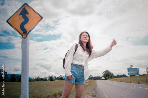 Billede på lærred Beautiful young asian woman with the backpack standing near a traffic sign and waits on an asphalt road in hot weather, A girl to catch a passing car for traveling