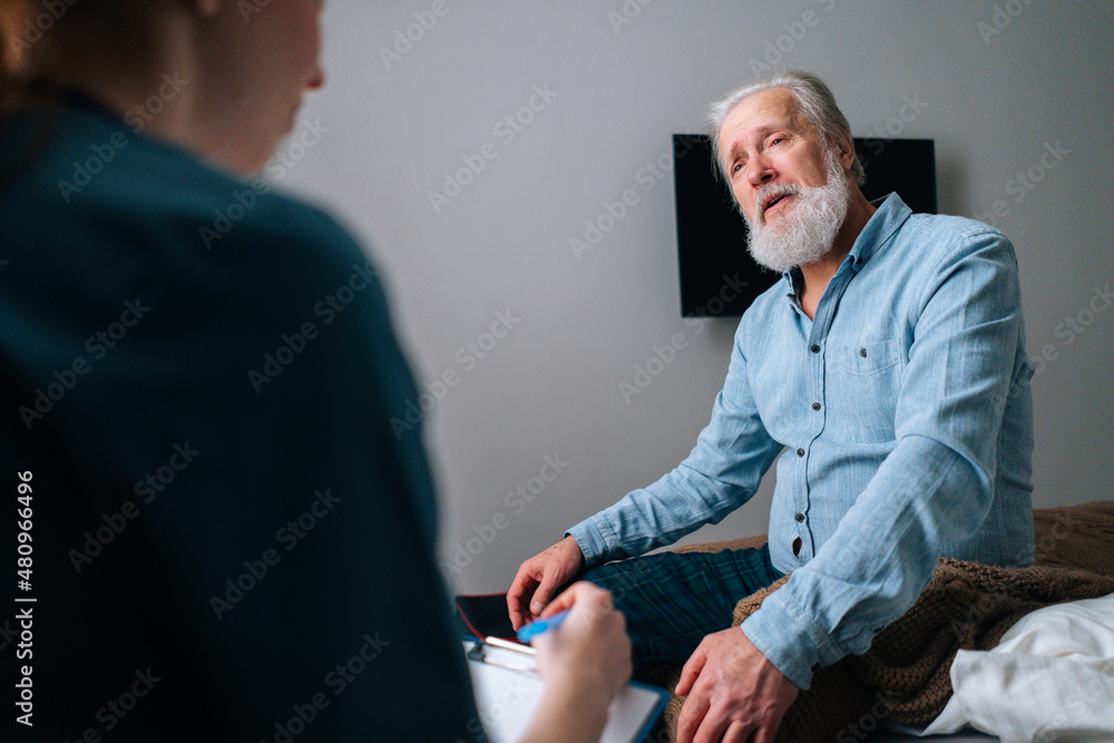 Obraz premium Close-up view from back of female doctor talking to illness senior male patient sitting on bed at nursing home, writing down diagnosis and symptoms in medical history on clipboard, selective focus.