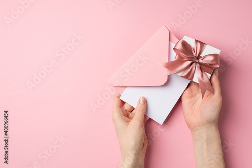 First person top view photo of st valentine's day decorations girl's hands holding small white giftbox with pink ribbon bow and envelope with letter on isolated pastel pink background with copyspace