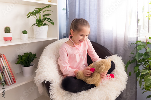 A preschool girl, dressed in a pink T-shirt, plays at home on an armchair in the hospital, listens to a teddy bear in a fanendoscope. photo