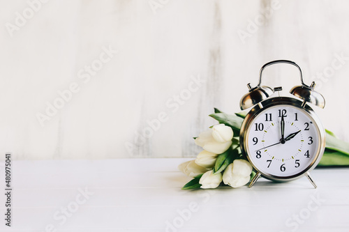 Daylight savings time concept. Set your clocks and to 2 am and spring ahead with this image of an alarm clock with white tulip flowers. Selective focus with blurred foreground and background with copy