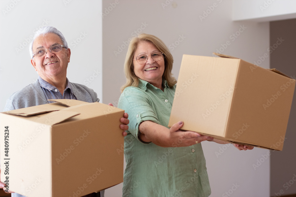 Thrilled elderly couple moving into new house. Retired husband and wife holding cardboard boxes, carrying things, smiling brightly. Real estate, purchase, moving concept Thrilled elderly couple moving into new house. Retired husband and wife holding cardboard boxes, carrying things, smiling brightly. Real estate, purchase, moving concept
