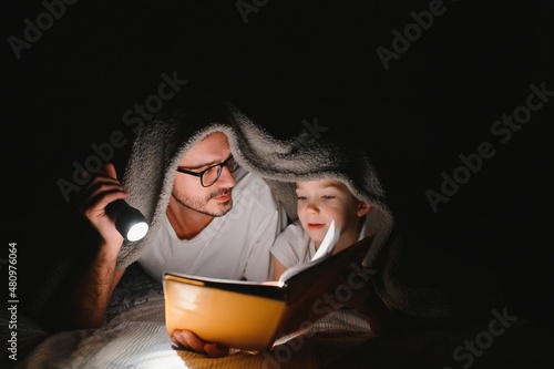 Happy family reading bedtime story under blanket in evening. Father and son spend time together. Father's Day.