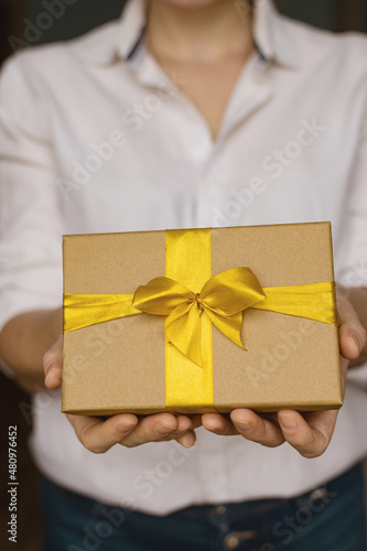 Fototapeta Naklejka Na Ścianę i Meble -  A young woman in a white shirt and jeans holds a gift for the holiday. Authentic dark photography. Gift greeting for holiday. Valentine's Day, Mother's Day, Women's Day, Birthday. Vertical