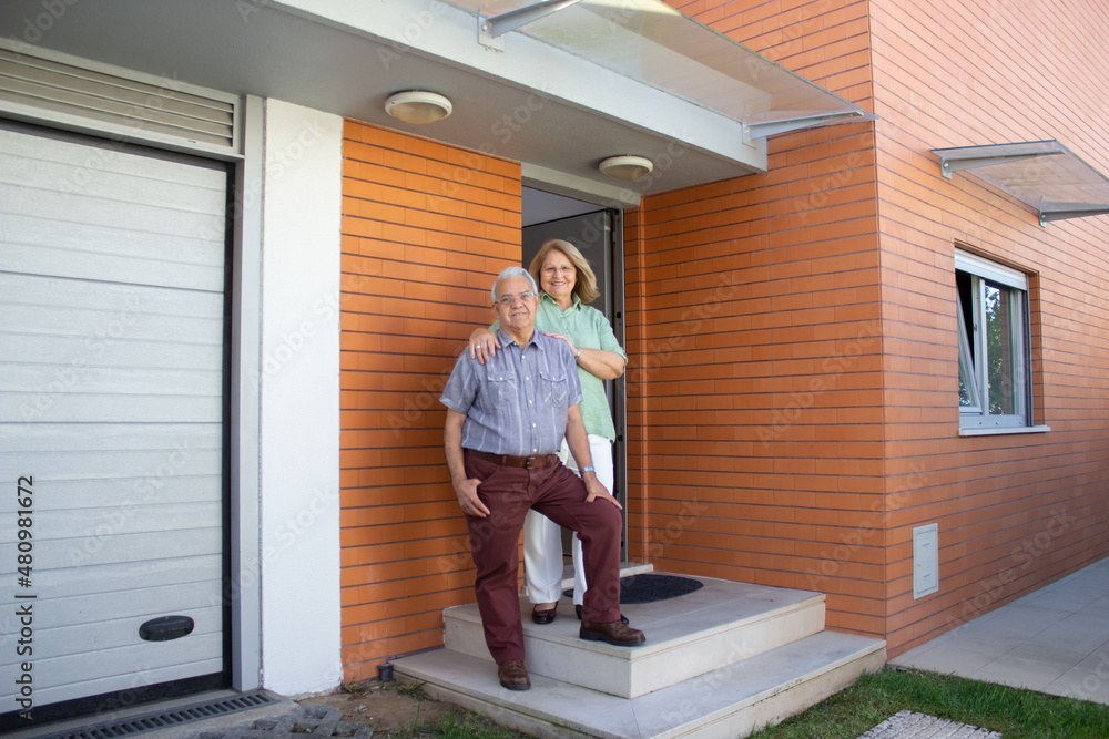 Happy retired family standing at entrance of new house. Elderly man and woman in casual clothes smiling at camera. Real estate, purchase, family concept