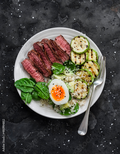 Balanced lunch - medium roast beef steak, grilled zucchini and couscous, avocado, spinach, egg salad on a dark background, top view