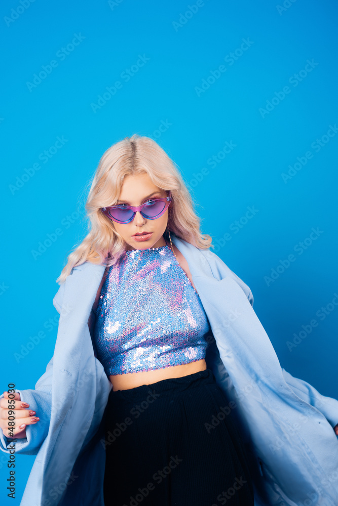 Obraz premium A girl in a blue jacket and purple glasses poses on a blue background and dances. Fashion photo in style 80s 90s party. Blonde girl in a shiny sweater and long earrings