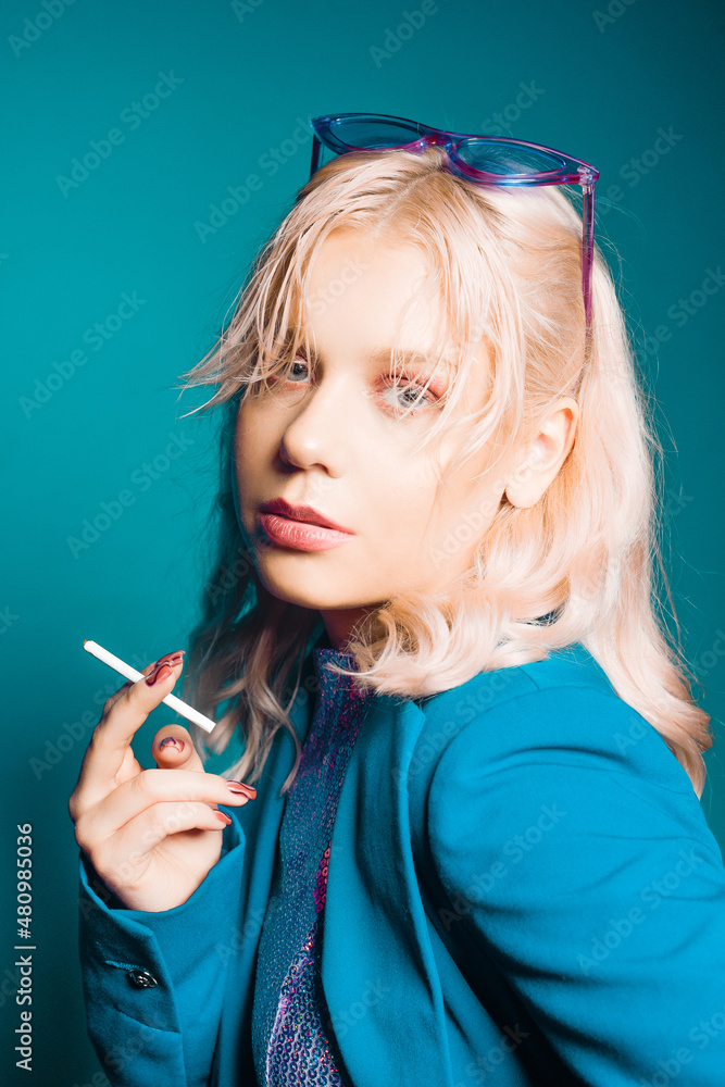 Obraz premium Incredibly fashionable blonde woman in a blue jacket and glasses in the style of 80s 90s poses. On a blue background. Fashionable woman smoking