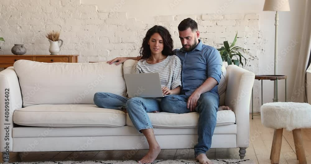 Beautiful millennial couple relaxing on couch with laptop, talking, discussing purchase on-line. Buying goods for house interior on internet web store, make order use website with electronic services Beautiful millennial couple relaxing on couch with laptop, talking, discussing purchase on-line. Buying goods for house interior on internet web store, make order use website with electronic services
