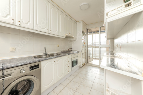 Long kitchen with white lacquered wood furniture, blue countertops and appliances with access to a glazed terrace
