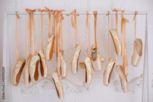 Many hanging ballet shoes on white wall background in studio. New pointe shoes with satin ribbons hanging on rank. Ballet shoes hang on bar in room. Concept of dance, ballet school, ballerinas clothes