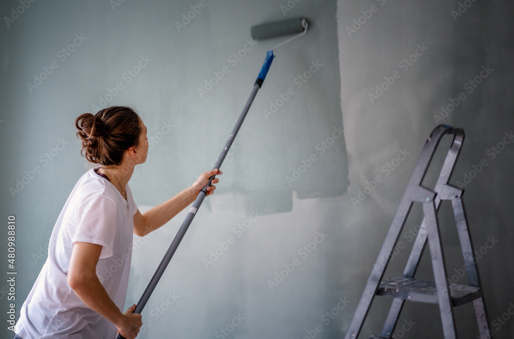 A young woman paints the walls in her house olive green using a roller. A young woman paints the walls in her house olive green using a roller.