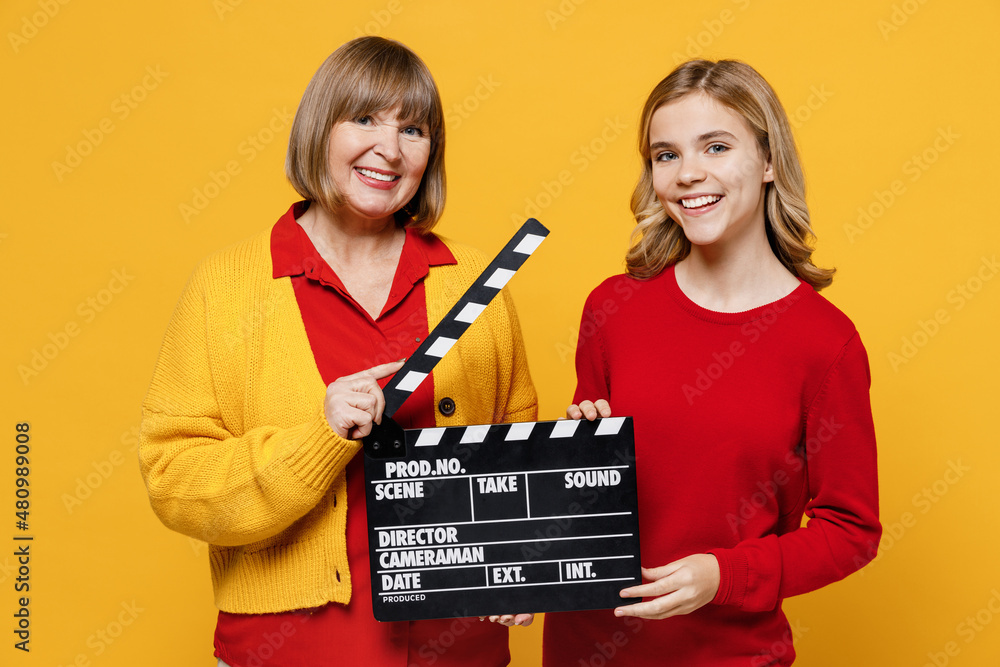 Happy fun woman 50s in red shirt with teenager girl 12-13 years old. Grandmother granddaughter hold classic black film make clapperboard isolated on plain yellow background. Family lifestyle concept.
