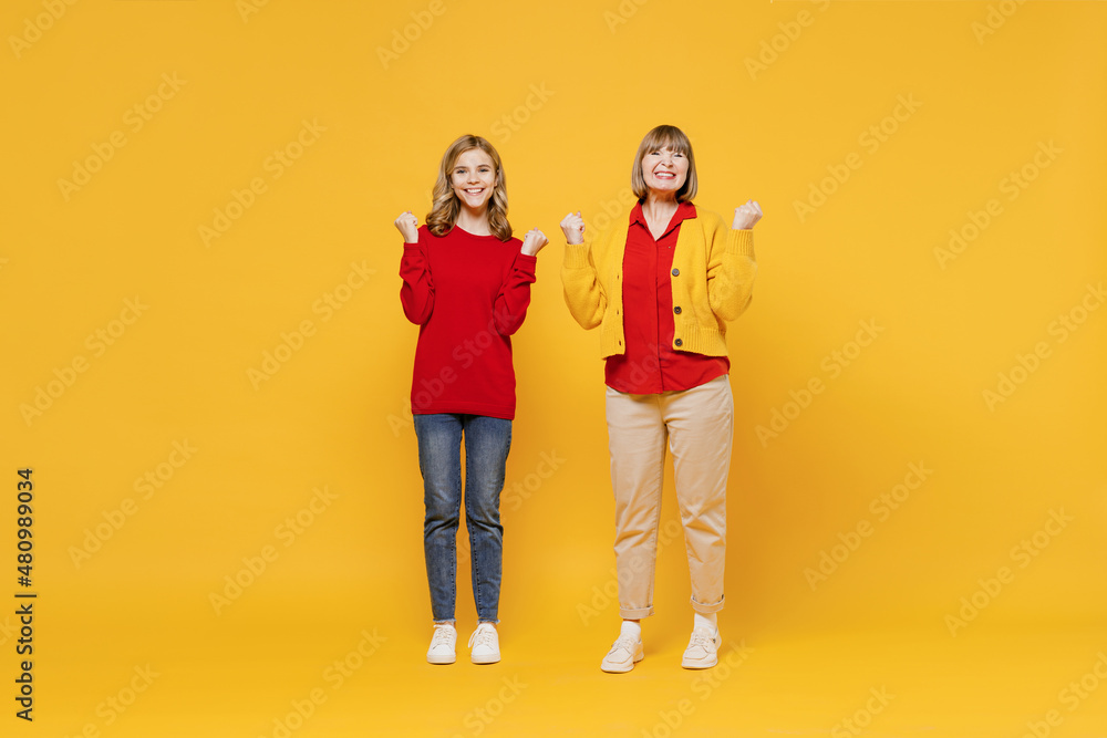 Full body cool woman 50s in red shirt have fun with teenager girl 12-13 years old. Grandmother granddaughter doing winner gesture celebrate clenching fists say yes isolated on plain yellow background