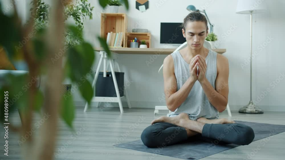 Slow motion of mixed race man meditating at home moving hands in namaste sitting on yoga mat in modern apartment. People and relaxation concept. Slow motion of mixed race man meditating at home moving hands in namaste sitting on yoga mat in modern apartment. People and relaxation concept.