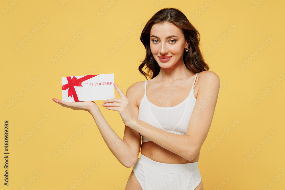 Smiling happy caucasian young brunette woman 20s in white underwear with perfect fit body hold gift certificate coupon voucher card for store isolated on plain yellow color background studio portrait. Smiling happy caucasian young brunette woman 20s in white underwear with perfect fit body hold gift certificate coupon voucher card for store isolated on plain yellow color background studio portrait.