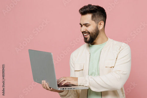 Young smiling happy cheerful caucasian man 20s wearing trendy jacket shirt hold use work on laptop pc computer isolated on plain pastel light pink background studio portrait. People lifestyle concept.