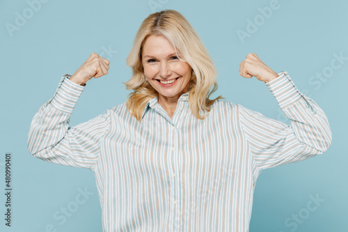 Elderly strong sporty woman 50s wear striped shirt showing biceps muscles on hand demonstrating strength power isolated on plain pastel light blue background studio portrait. People lifestyle concept.
